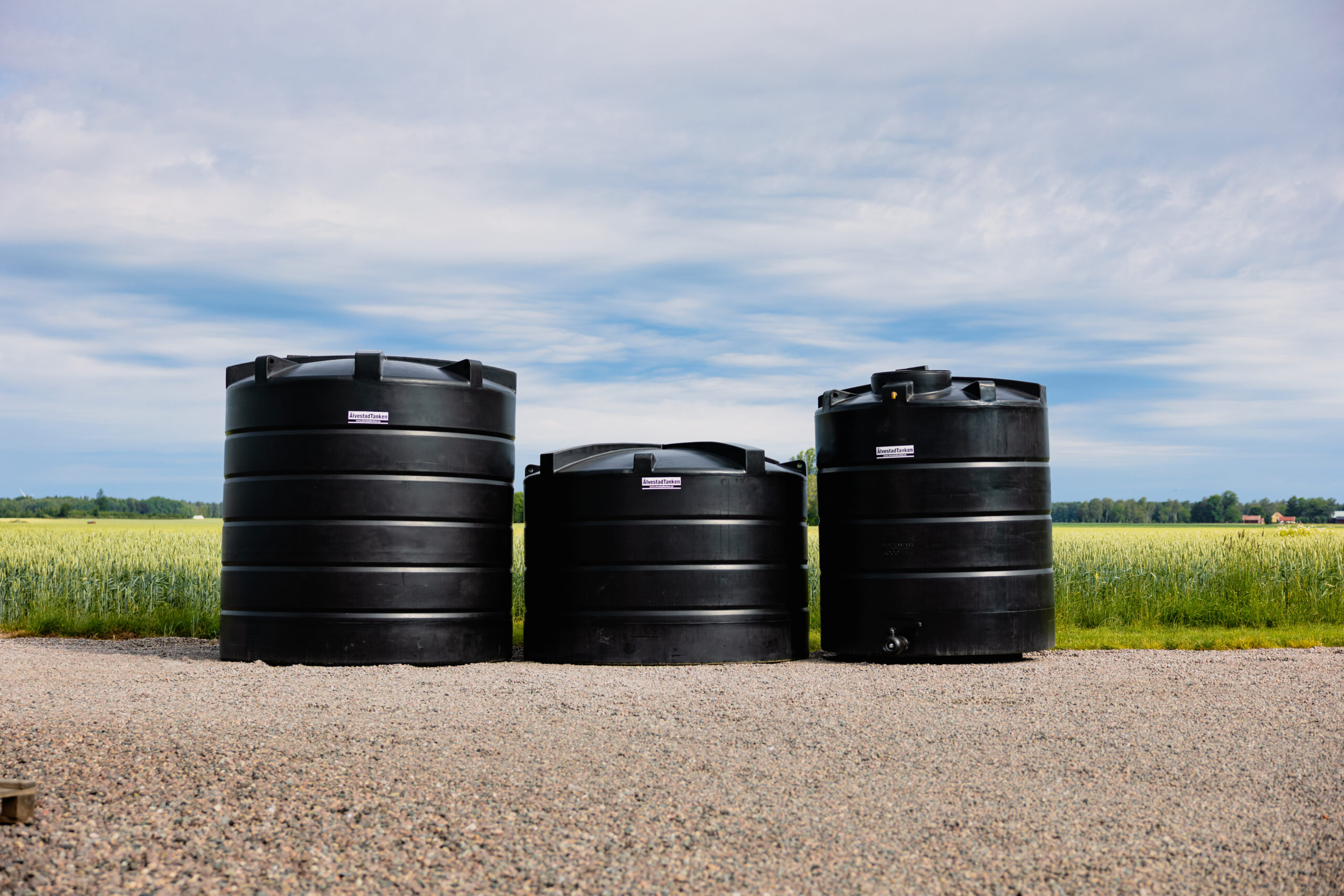 Tre stora vattentankar i svart plast står på en grusyta, med gröna fält och en molnig himmel i bakgrunden.
