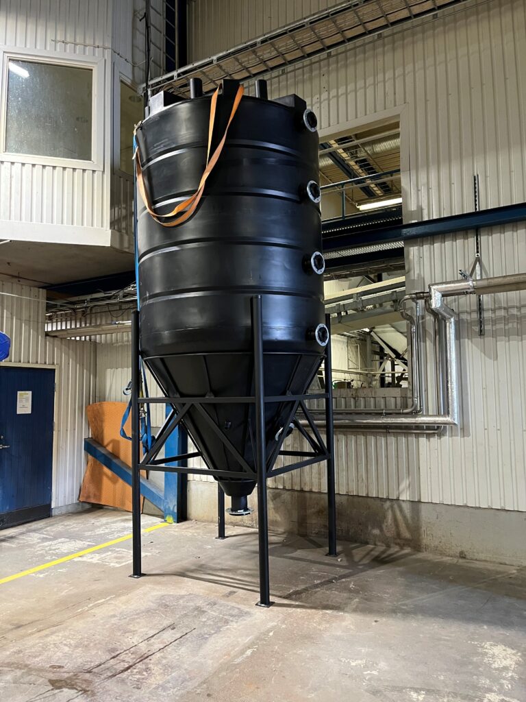 En stor svart industriell lagringstank med flera röranslutningar står på metallben inne i en fabrik med vita panelväggar och synliga rör. En orange lyftstropp är fäst över tanken.