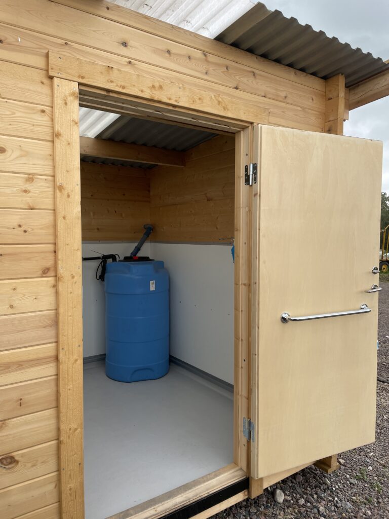I ett träskjul med öppen dörr finns en blå vattentunna av plast och en svart pump. Innerväggarna är av trä och golvet är ljusgrått. Skjulet har ett tak av korrugerad plast.