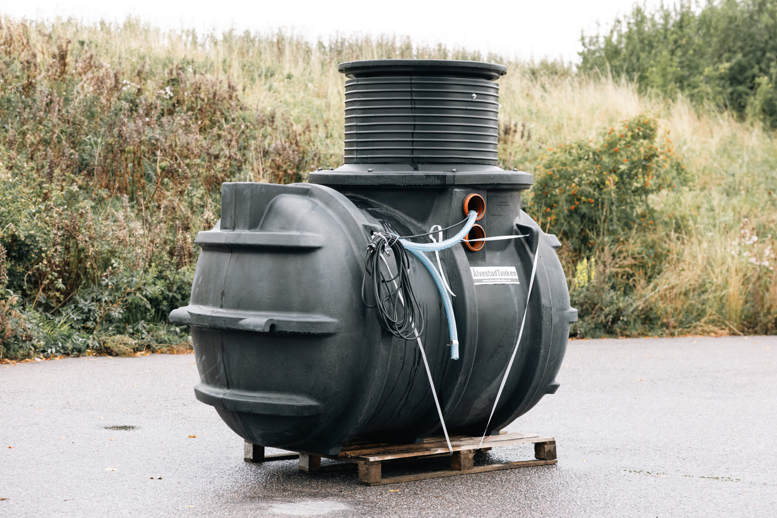 En stor svart plasttank med rör och kablar står på en träpall utomhus, med gräs och buskar i bakgrunden.