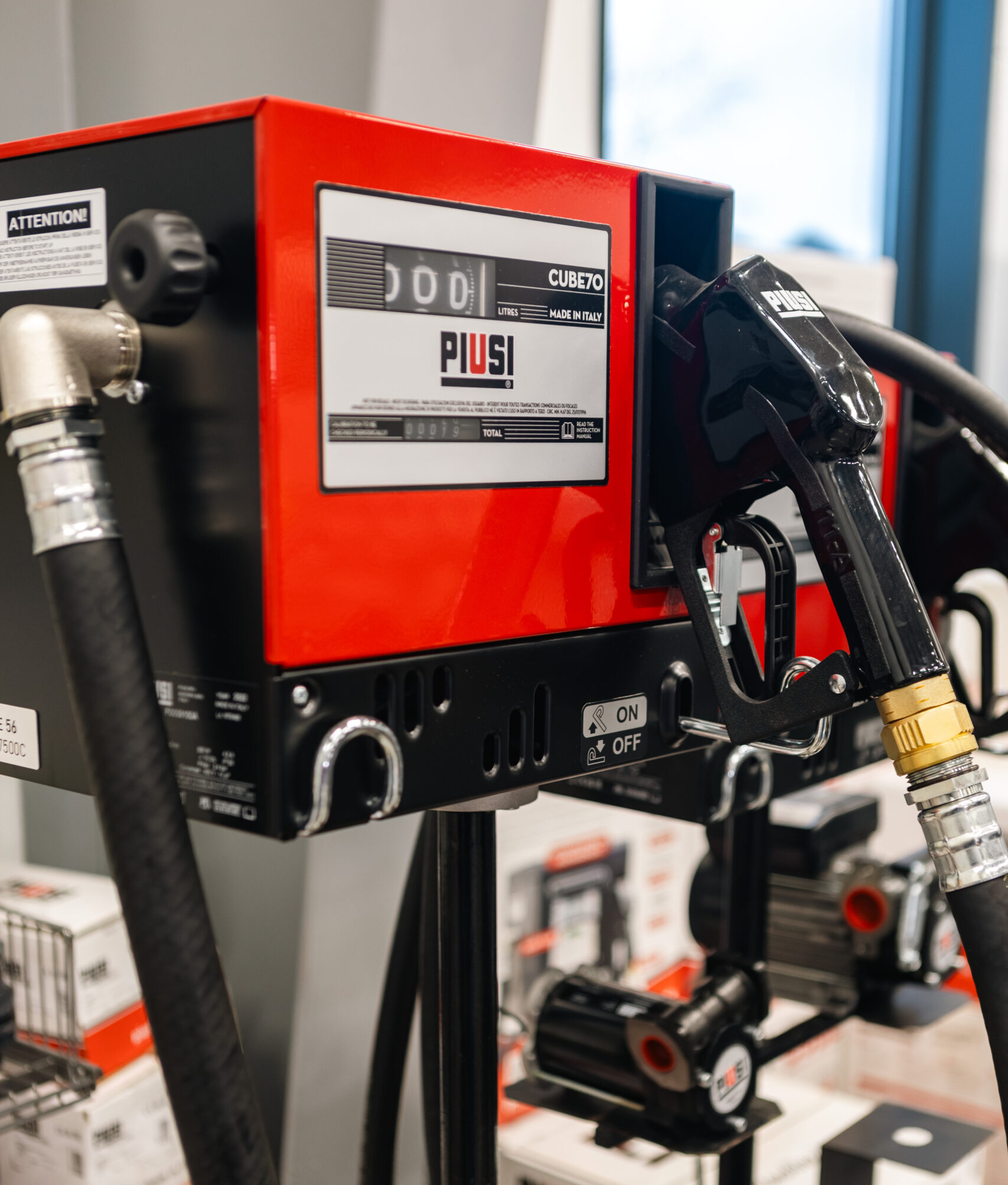 Närbild av en röd och svart bränslepump märkt "PIUSI" med slangar och munstycke, placerad inomhus i närheten av produktlådor och andra pumpenheter.