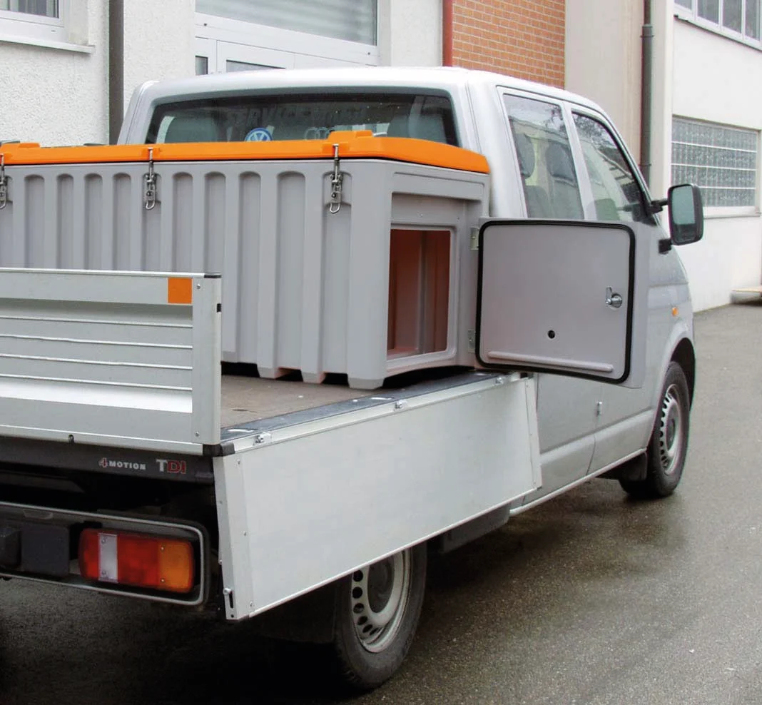 En liten vit pickup med öppet sidoutrymme och en förvaringslåda av typen CEMbox 750 liter med sidodörr – med grå kaross och orange lock – fastspänd på flaket, parkerad på en gata i stan nära en byggnad.