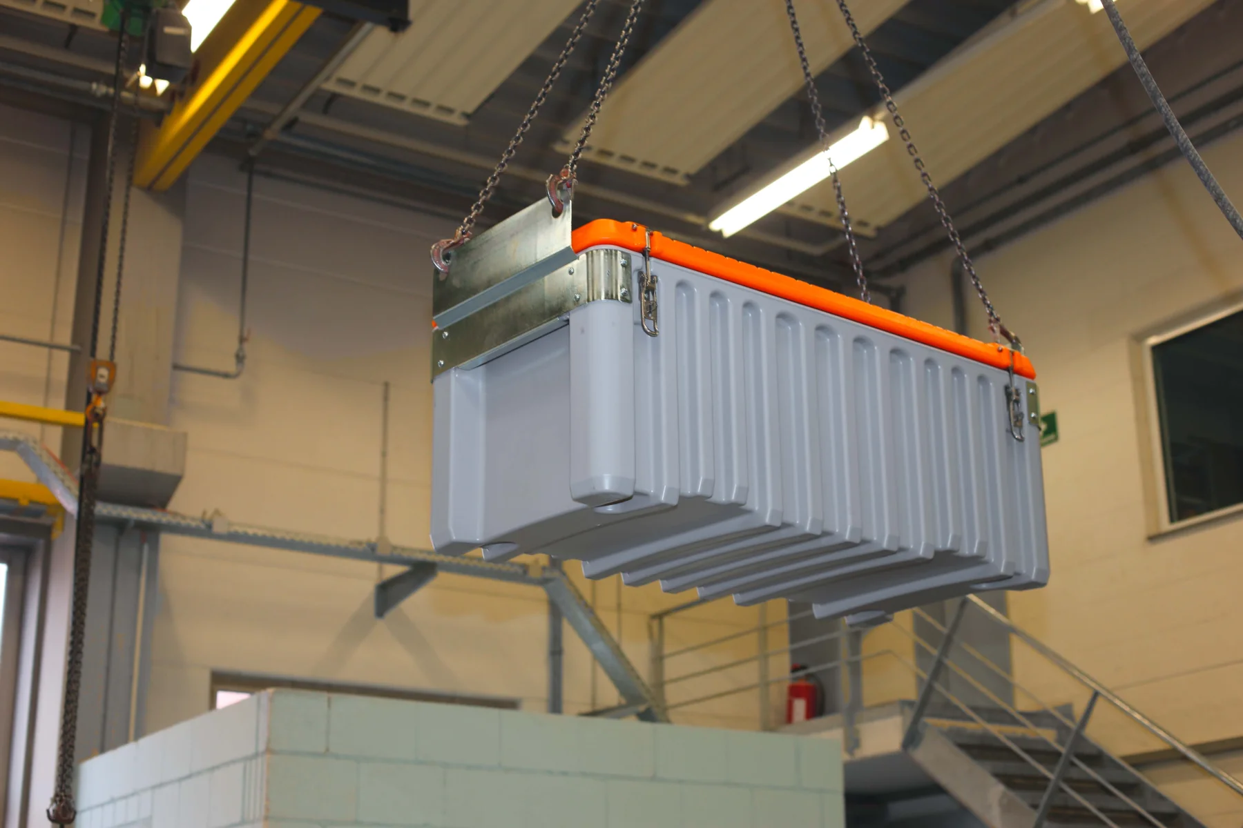 Ett stort grått industriellt batteripaket hänger i kedjor i en fabriksmiljö, med metallbalkar och trappor synliga i bakgrunden.