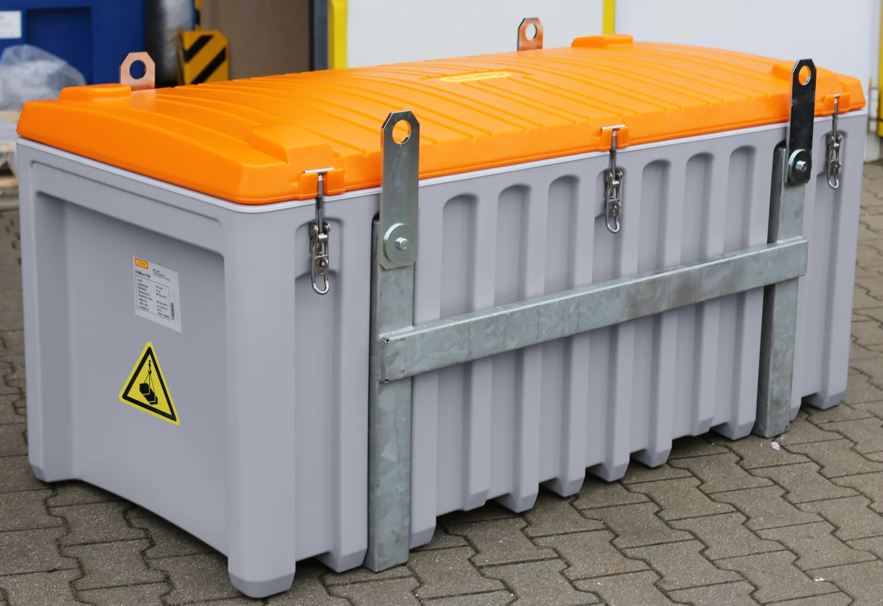 En stor grå behållare med ett orange lock, metallfästen och varningsetiketter står på en asfalterad yta intill en byggnadsvägg.