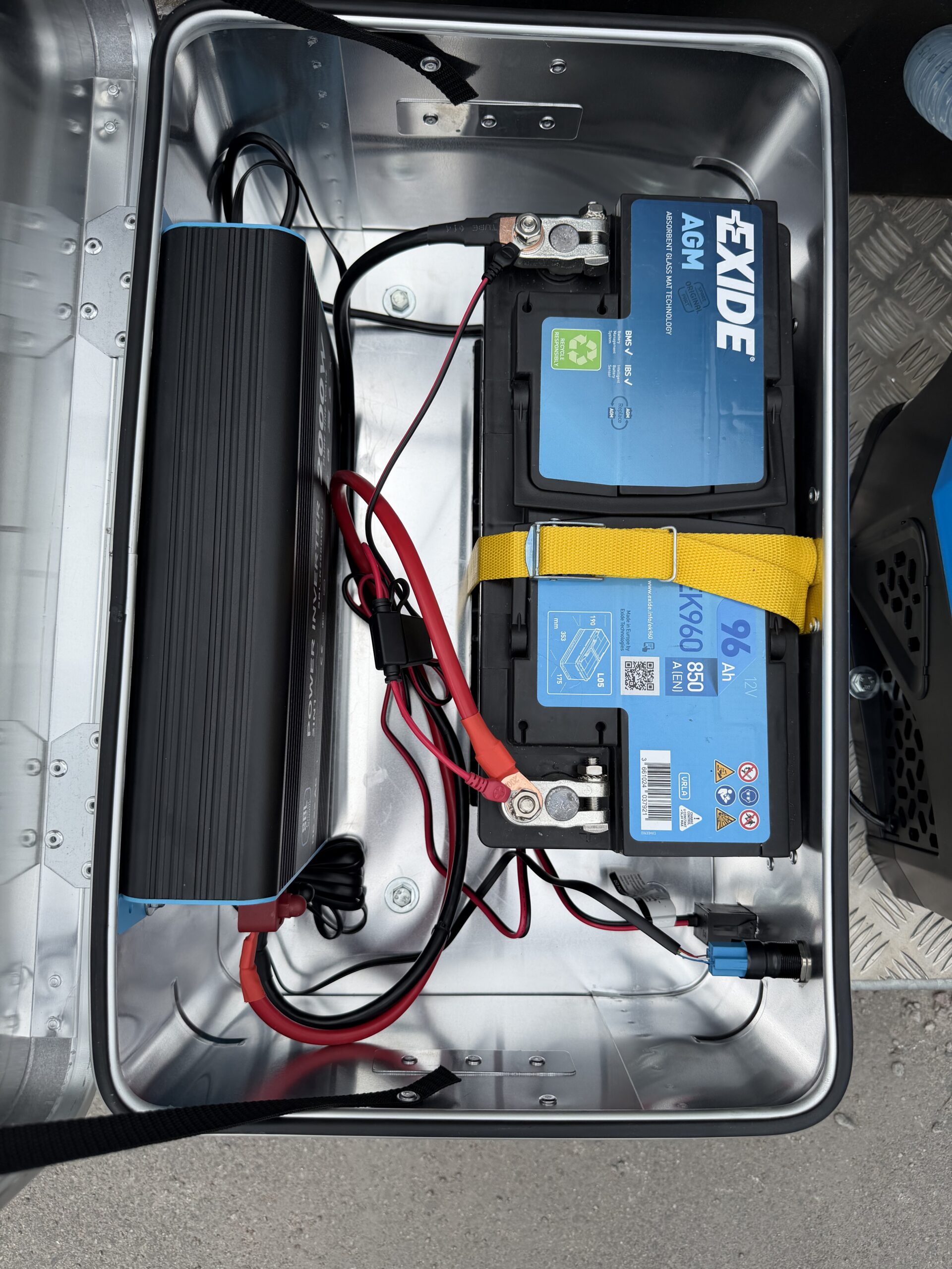 En öppen metallåda som innehåller två Exide AGM-batterier, strömkablar, ett gult band som håller fast batterierna och en elektronisk enhet, där alla delar är anslutna inuti. Uppställningen ingår i Bevattningsplattform 700 liter och är placerad på en grå yta.