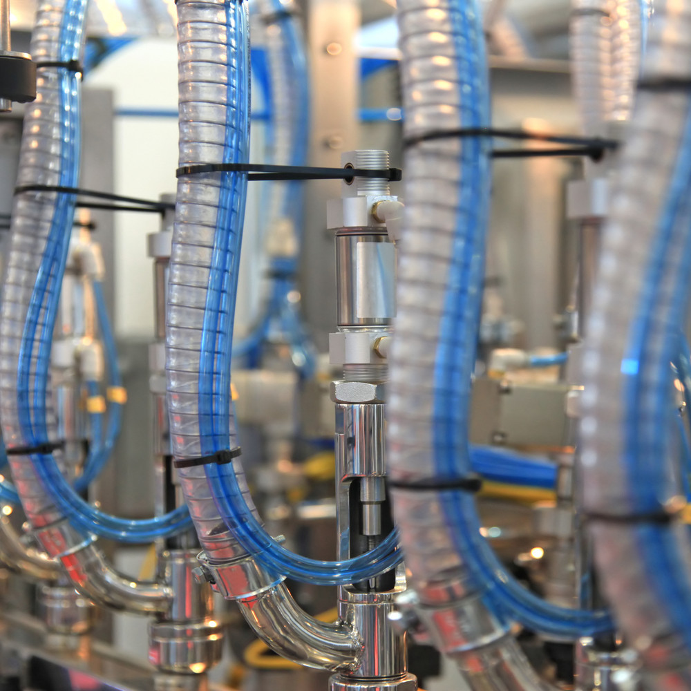 Närbild på Vattenslang Metalflex-slangar av livsmedelsgodkänd blå och genomskinlig plast, anslutna till metallmaskiner, troligen använda i en industri- eller tillverkningsprocess. Fokus ligger på slangarna och kopplingarna, medan utrustningen i bakgrunden är mjukt suddig.