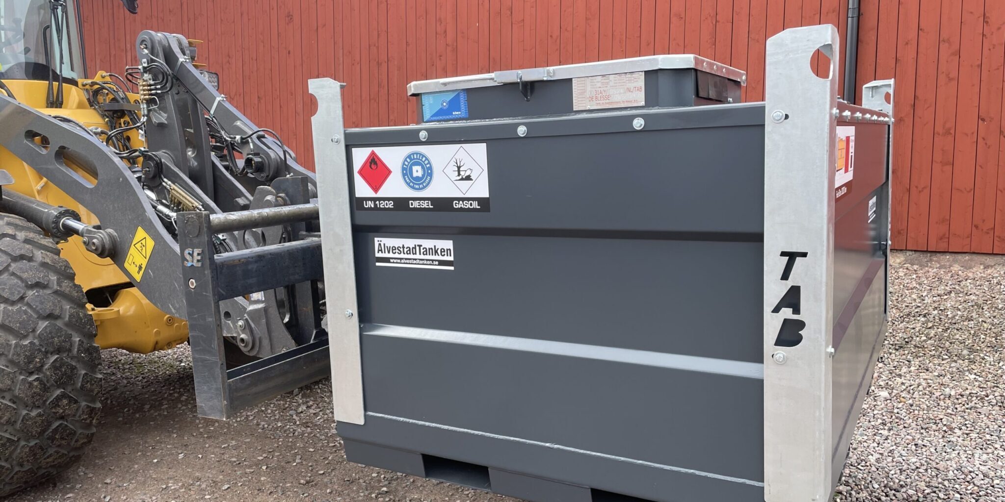 En gaffeltruck kopplas till en stor, grå Fuel Box ADR dieseltank - 450-3000 liter - med etiketter för fara och brandfarlig vätska, framför en röd träbyggnad på en grusyta. Vi lanserar nu Fuel Box ADR dieseltankar!.