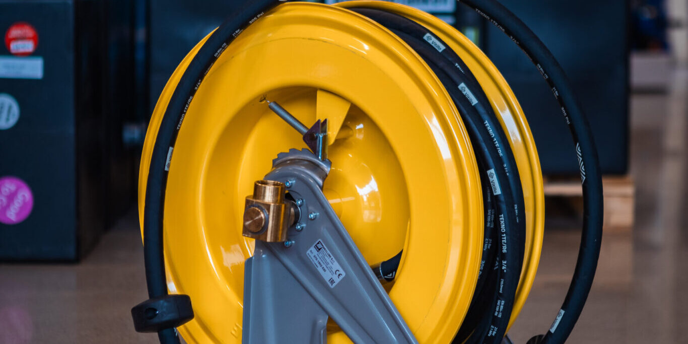 En gul industrislangvinda med svart slang, en del av vårt professionella slangvindor-sortiment, visas på ett polerat golv i en verkstad, med verktyg och utrustning som syns i den suddiga bakgrunden.